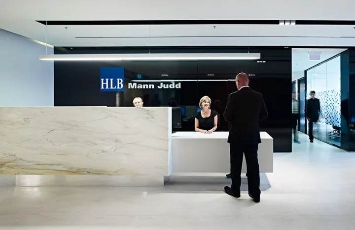 A modern office reception area with two receptionists behind a marble desk and a man in a suit standing in front, reflecting HLB Mann Judd’s commitment to sustainable growth. Another person walks through a glass doorway in the background.