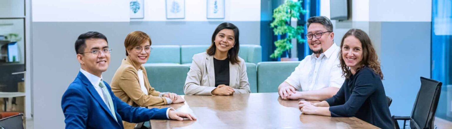 Five people in business attire sit around a conference table, smiling at the camera in a modern office setting with plants and framed art on the walls. - Emapta
