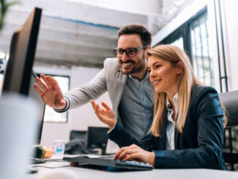 Two colleagues in business attire smiling and looking at a computer monitor in a modern office. One person is seated, typing, while the other stands beside, gesturing towards the screen.