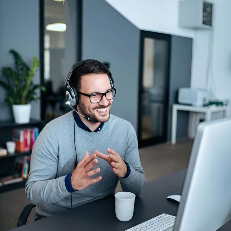 A man wearing glasses and a headset sits at a desk, smiling and gesturing during a video call. A coffee cup is in front of him, and office furniture and plants are visible in the background
