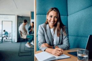 A woman in a gray blazer sits at a desk with an open notebook, smiling at the camera. A laptop and a glass of water are on the table, and two people are blurred in the background.