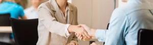Two people in business attire shaking hands across a desk in an office setting, suggesting an accountant hiring interview or meeting.