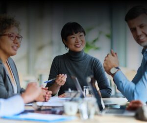 Four people sit at a table engaged in a business meeting, with documents, laptops, and pens visible; one woman in the center is smiling.