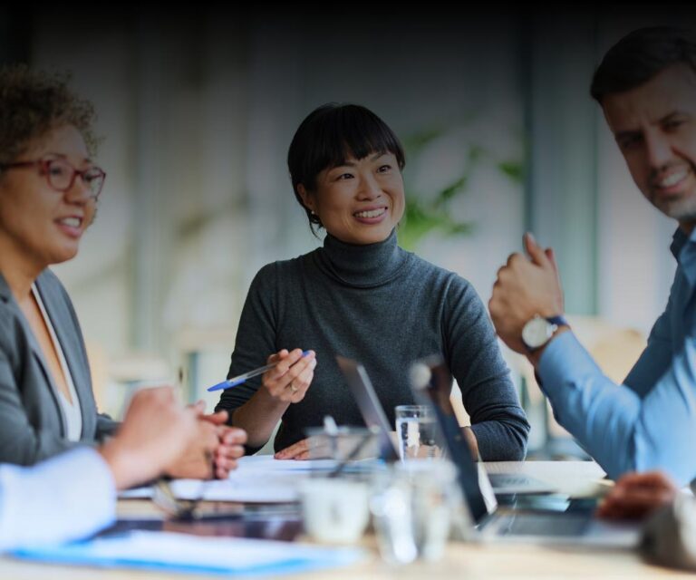 Four people sit at a table engaged in a business meeting, with documents, laptops, and pens visible; one woman in the center is smiling.