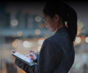 A person in a suit using a tablet and wearing a headset, standing against a blurred city background with lights.