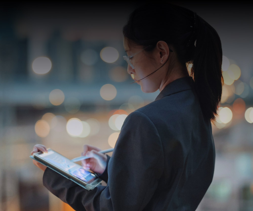 A person in a suit using a tablet and wearing a headset, standing against a blurred city background with lights.