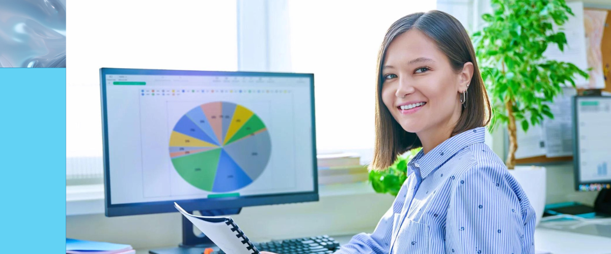 A woman sits at a desk holding documents and smiling at the camera, with a computer monitor behind her displaying a colorful pie chart—reflecting efficient data entry processes. There are plants and office supplies in the background.