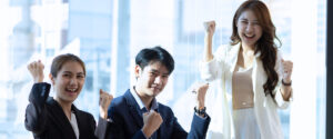 Three business professionals in formal attire stand indoors, smiling and raising their fists in a gesture of celebration or success.