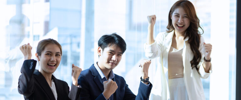 Three business professionals in formal attire stand indoors, smiling and raising their fists in a gesture of celebration or success.