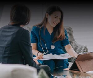 A healthcare professional with a stethoscope reviews documents and a tablet with another person sitting across a glass table.