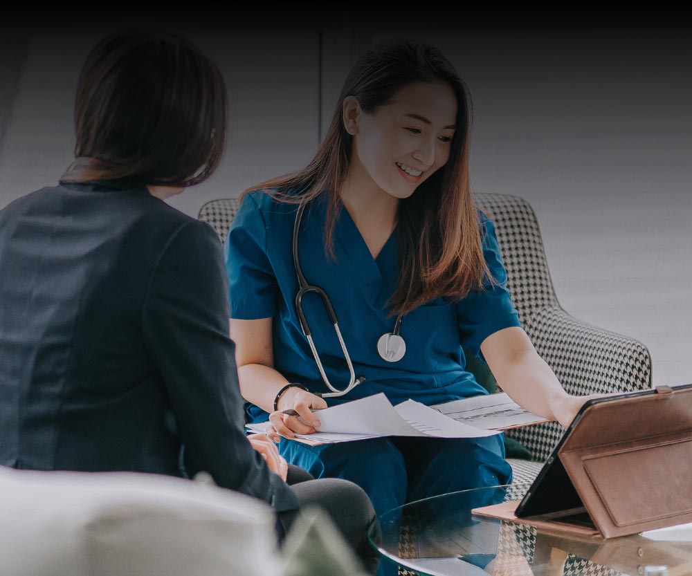A healthcare professional with a stethoscope reviews documents and a tablet with another person sitting across a glass table.