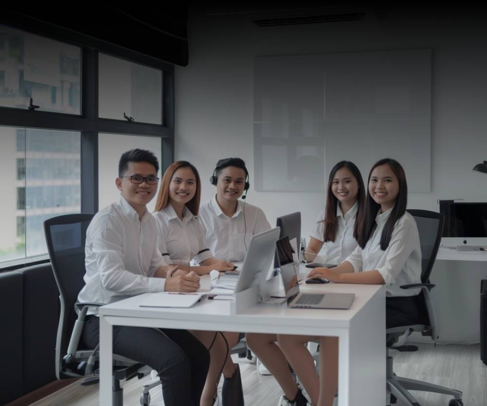 Five people in business attire sit around a table in a modern office, smiling at the camera. This high-performing customer experience team is ready to build success, with laptops and notebooks spread out and large windows in the background.