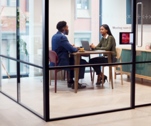 Two people sit across from each other in a modern glass-walled meeting room, discussing differences between subcontracting and outsourcing. One has a laptop open, and a sign says Meeting Room 8.