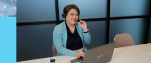 A woman wearing a headset smiles while sitting at a desk with a laptop, appearing to be in an Outsourced Customer Support or customer service video call in a modern office environment.