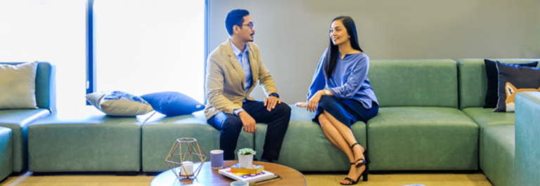 A man and a woman sit on a green couch in a modern office lounge, discussing the cost of living in the Philippines. A coffee table with books, a plant, and decor is in front of them against a minimal, uncluttered background.