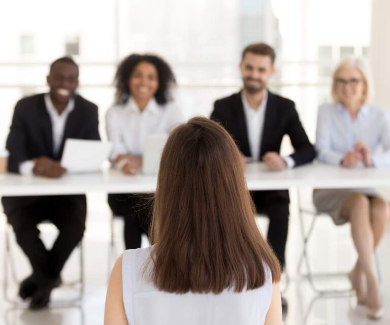A woman with brown hair sits facing four people in business attire, who are discussing must have skills and how virtual assistant skills can improve your business during a job interview in a bright, modern office setting.
