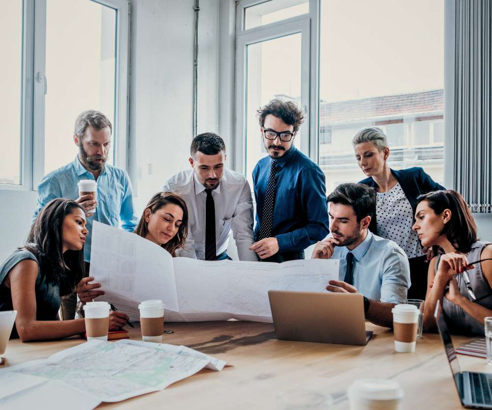 A group of eight professionals gathered around a table, examining large blueprints and discussing supply chain management. Laptops, paper cups, and documents are spread out on the table in a bright office setting.