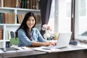 A woman sits at a desk using a laptop, smiling at the camera. She is in a bright room with shelves of books, papers, and flowers—a comfortable workspace where bulletproof data security keeps her information safe.