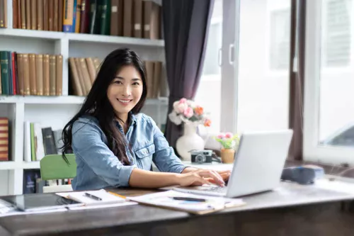 A woman sits at a desk using a laptop, smiling at the camera. She is in a bright room with shelves of books, papers, and flowers—a comfortable workspace where bulletproof data security keeps her information safe.