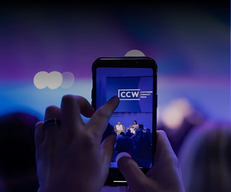 A person captures a photo of the stage with a smartphone at CCW Las Vegas 2025. The phone screen shows the vibrant stage and audience, bathed in purple and blue lighting—perfect for a Recap of this New Era Insights event.
