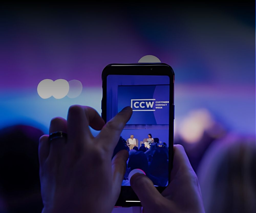 A person captures a photo of the stage with a smartphone at CCW Las Vegas 2025. The phone screen shows the vibrant stage and audience, bathed in purple and blue lighting—perfect for a Recap of this New Era Insights event.