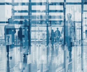 Abstract image with a blue tint showing silhouettes of people walking through a modern building with glass walls, reflections, and overlapping layers, creating a sense of motion and transparency.