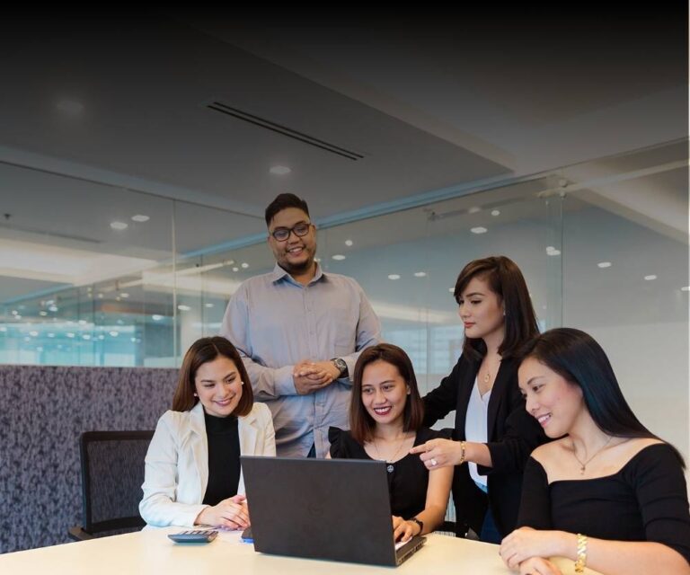 Five people in business attire are gathered around a laptop in a modern office with glass walls, smiling and engaging in a collaborative discussion on managing high turnover.