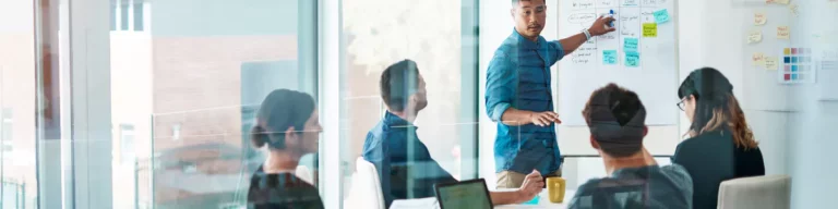 A man stands at a whiteboard covered with notes, presenting strategies for effective outsourcing to four colleagues seated at a meeting table in a modern glass-walled office.