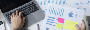 A person working on a laptop while holding a financial report with bar and pie charts, surrounded by additional printed graphs and a calculator—perfect for reviewing Outsourcing FAQs or other frequently asked questions about financial processes.