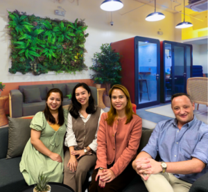 Four people sit on a sofa in a modern office lounge with greenery, cozy seating, and glass meeting pods. Smiling and relaxed, they discuss the 2026 outsourcing guide and why so many businesses outsource to the Philippines.