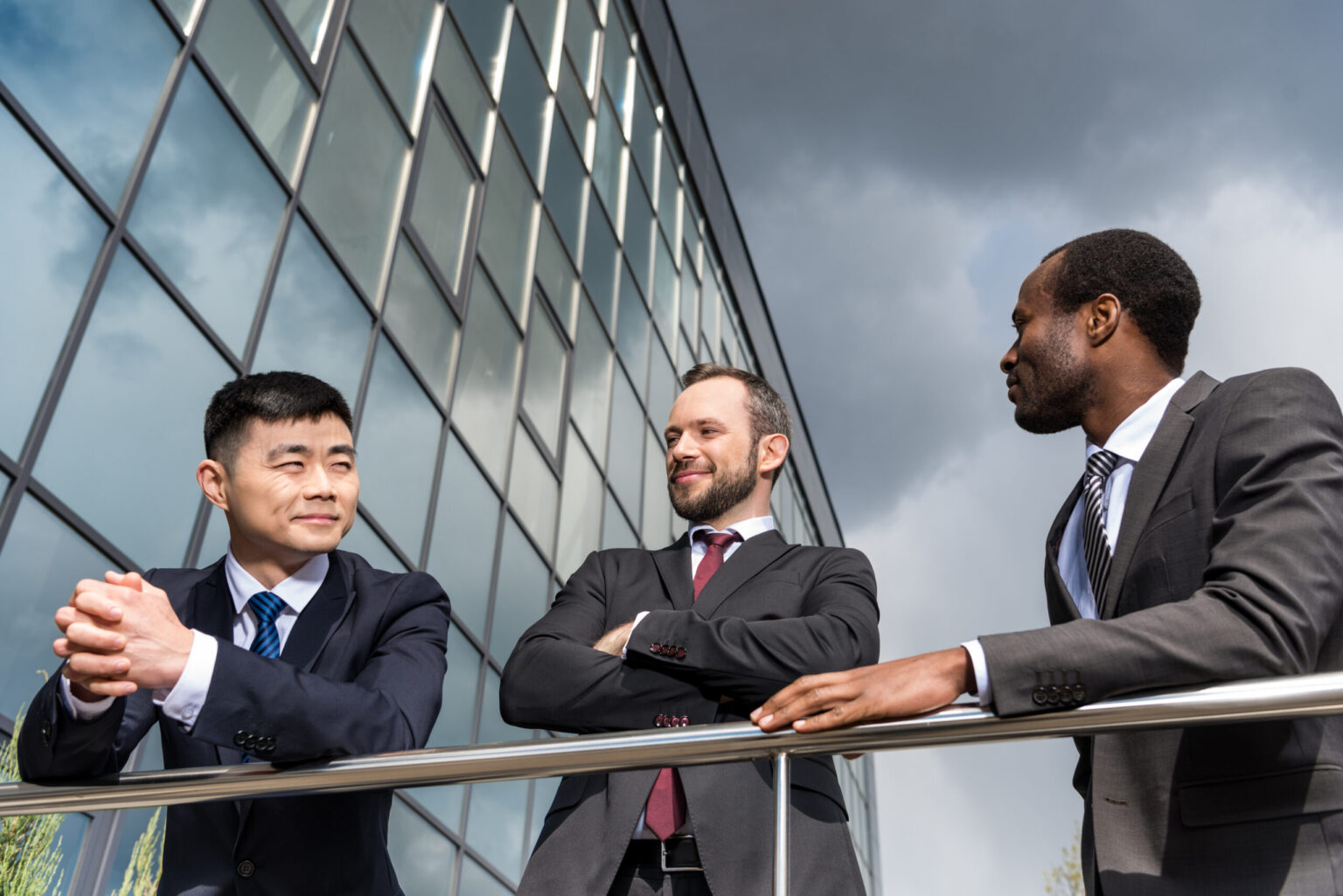 Three men in business suits stand and converse outside a modern glass building under dark clouds, appearing relaxed and engaged in a discussion about US companies outsourcing operations.