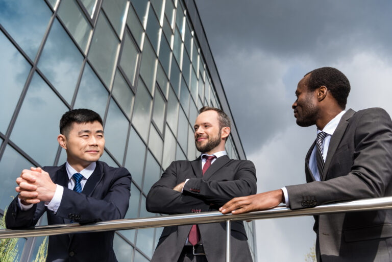 Three men in business suits stand and converse outside a modern glass building under dark clouds, appearing relaxed and engaged in a discussion about US companies outsourcing operations.