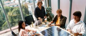 Four people collaborate around a table with a large solar panel, laptops, and documents, addressing business-critical demands in a bright office with large windows and sunlight streaming in.