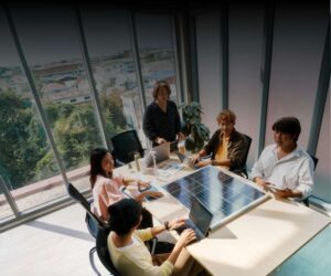 Five people sit around a conference table in a bright office with large windows, discussing business-critical demands in solar panels and working on laptops and tablets. A solar panel is placed in the center of the table.