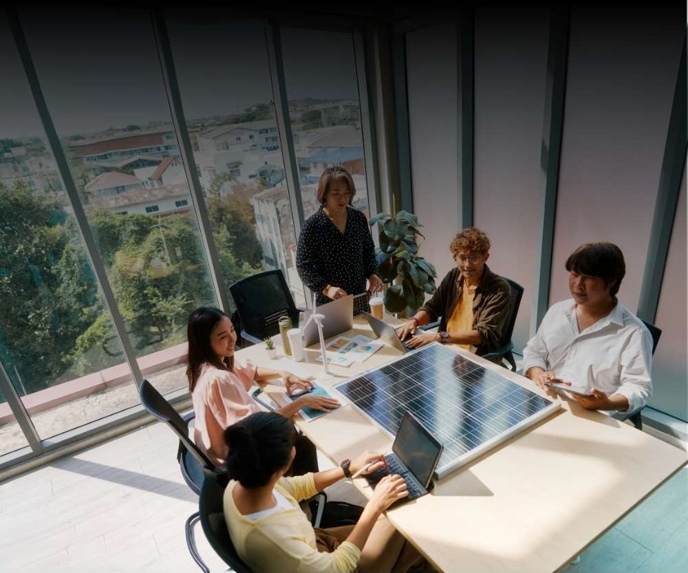 Five people sit around a conference table in a bright office with large windows, discussing business-critical demands in solar panels and working on laptops and tablets. A solar panel is placed in the center of the table.