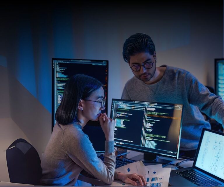 Two people in a dimly lit office work together at a desk with multiple monitors displaying code, supporting a 24/7 operation. One sits thinking, while the other stands and points at a screen. Papers and a laptop are spread out on the desk.