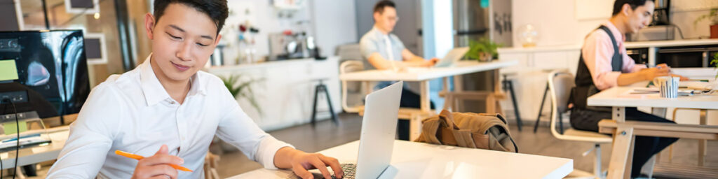 Three people working at desks in a modern, open office space. One person in the foreground is writing and using a laptop.