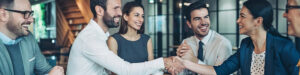 A group of business professionals sit around a table, smiling and laughing. Two people in the center are shaking hands, celebrating the benefits of compliance outsourcing in a modern office setting.