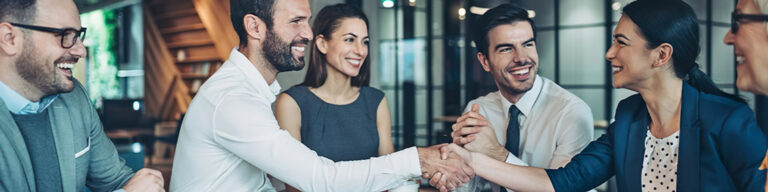 A group of business professionals sit around a table, smiling and laughing. Two people in the center are shaking hands, celebrating the benefits of compliance outsourcing in a modern office setting.