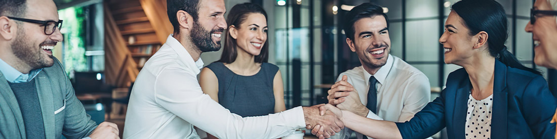 A group of business professionals sit around a table, smiling and laughing. Two people in the center are shaking hands, celebrating the benefits of compliance outsourcing in a modern office setting.