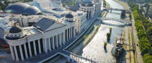 Aerial view of neoclassical buildings, a domed museum, and a stone bridge along the Vardar River in Skopje, North Macedonia—an emerging nearshoring hub for European businesses by 2026—with a replica ship and tree-lined riverbanks.