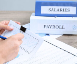 A person reviews a financial chart with a pen at a desk. In the background, two binders labeled SALARIES and PAYROLL are stacked, highlighting Payroll Outsourcing Statistics in an organized office environment.