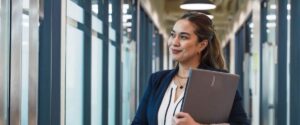 A woman in business attire stands in a hallway holding a closed laptop, smiling and looking out a window—reflecting on the differences accountants face between the US, UK, Australia, and the Philippines.