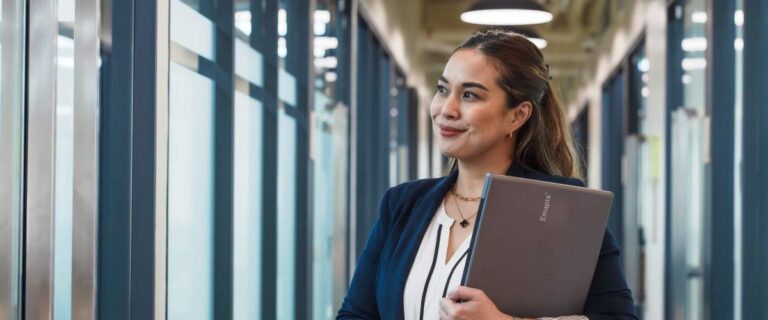 A woman in business attire stands in a hallway holding a closed laptop, smiling and looking out a window—reflecting on the differences accountants face between the US, UK, Australia, and the Philippines.