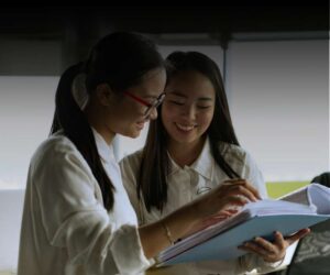 Two young women in white shirts stand together, smiling as they review paperwork or a large open book in a brightly lit indoor setting, discussing industry trends and strategies for business growth.