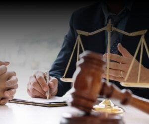 Close-up of two people in suits at a desk with a gavel, scales of justice, and clipboard, suggesting legal research or law firms in discussion. One person writes while the other gestures during this professional exchange.