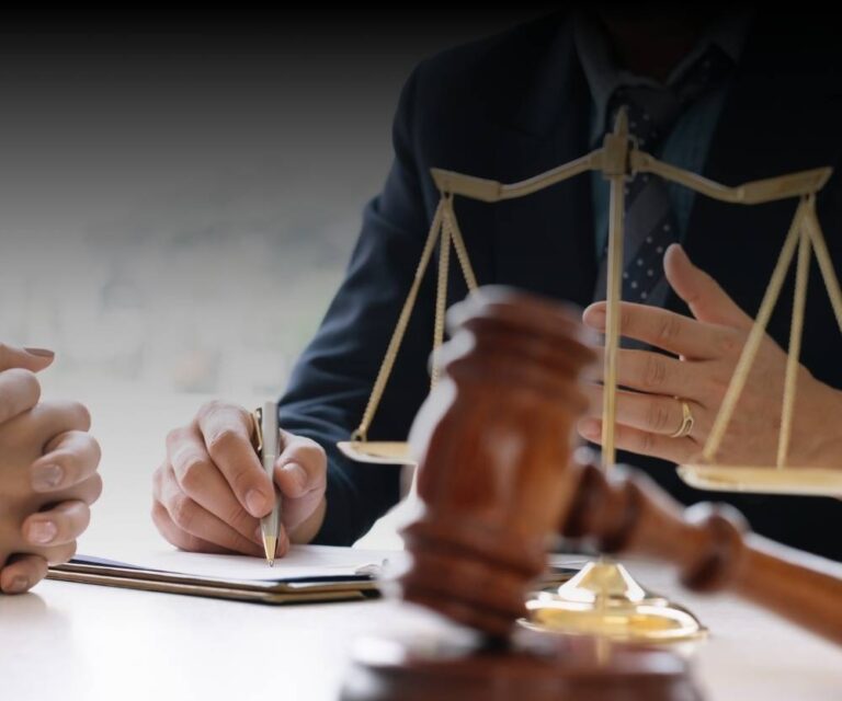 Close-up of two people in suits at a desk with a gavel, scales of justice, and clipboard, suggesting legal research or law firms in discussion. One person writes while the other gestures during this professional exchange.