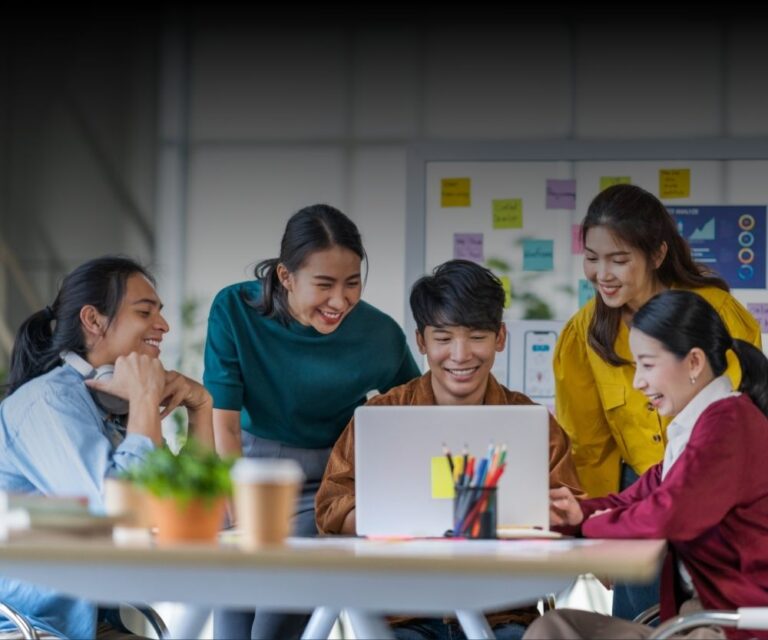 Five young adults gather around a table, smiling and looking at a laptop screen as they discuss outsourcing options. The group appears engaged and collaborative, with colorful sticky notes and office supplies in the background.