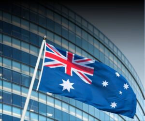 The Australian flag waves on a pole in front of a modern glass building, home to an Emapta offshore partner, showing the Union Jack and white stars on a blue background.