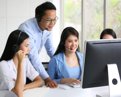 Three women and one man, all wearing headsets, work together at a computer in a bright office. The man stands behind the women, smiling and providing guidance as they look at the monitor.